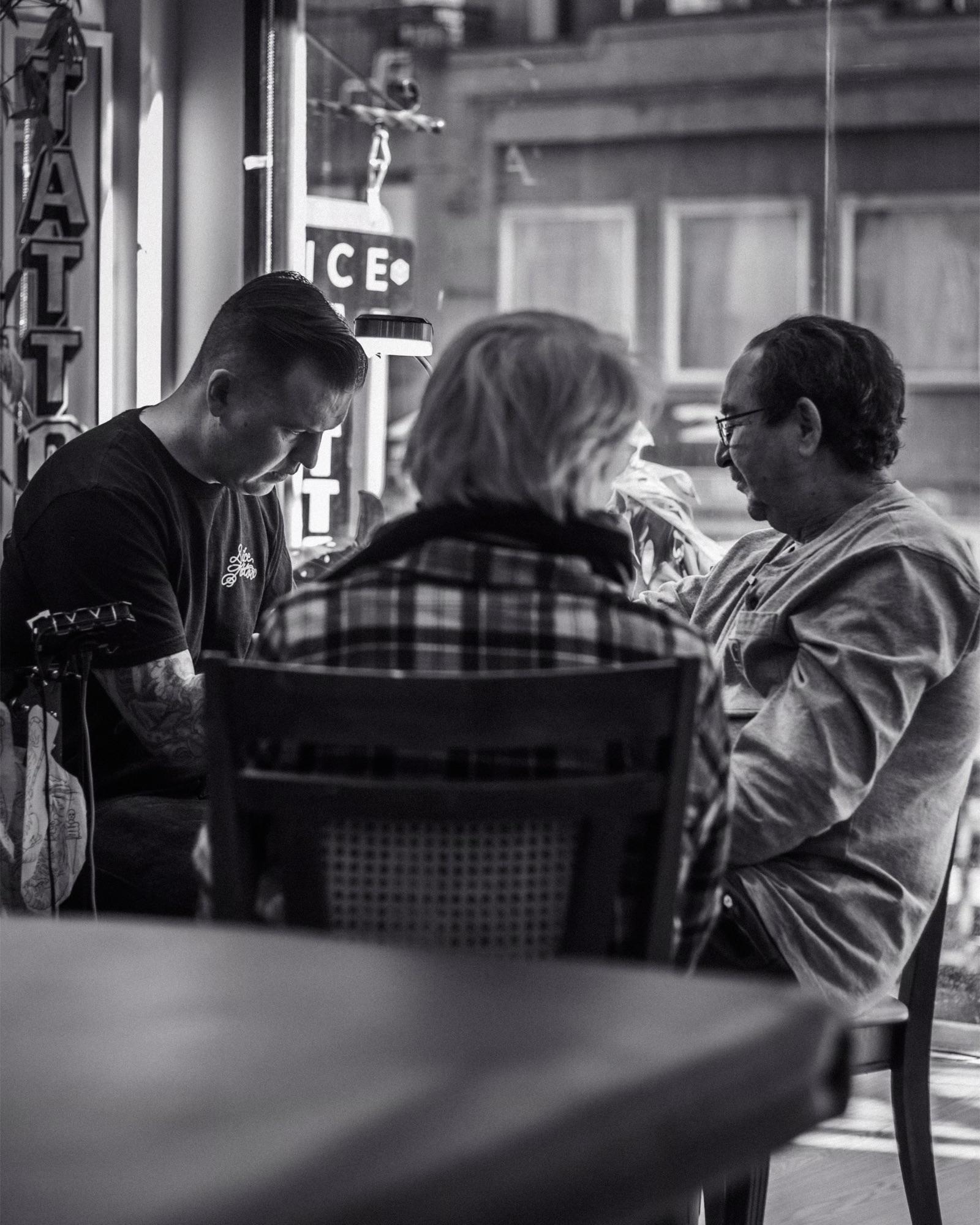Mike Stapleton tattooing at a table by the shop window while two people chat