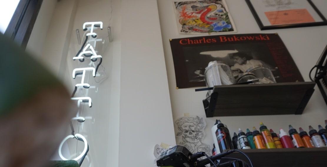 Studio wall with a neon tattoo sign and a Charles Bukowski poster